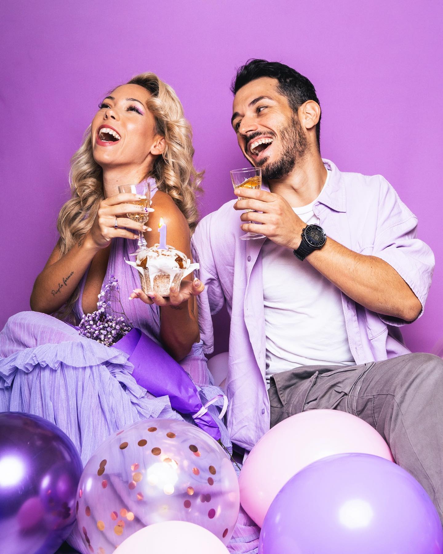 Pareja riendo y celebrando con bebidas y un cupcake en un ambiente festivo con globos.
