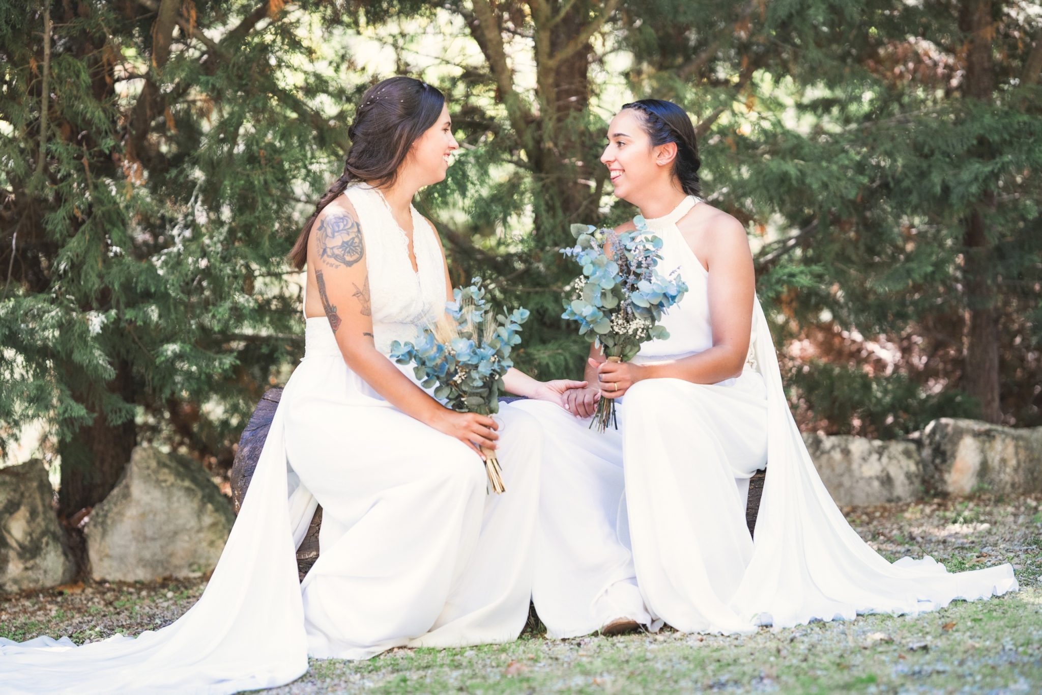 Dos novias sentadas una al lado de la otra, sosteniendo ramos y compartiendo una mirada afectuosa.