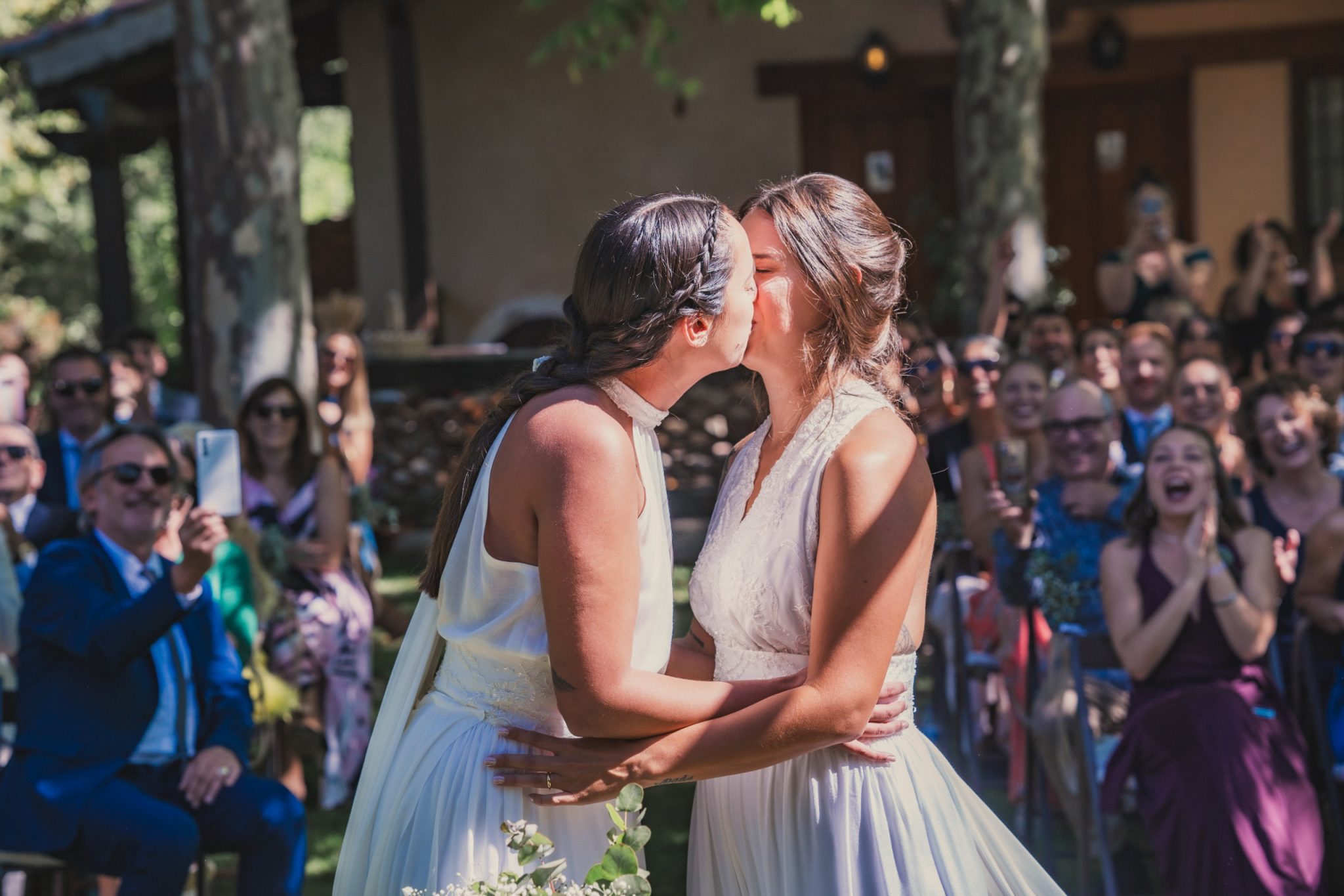 Dos novias se besan mientras los invitados celebran en su boda.
